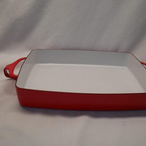 Puede incluir: Una fuente de horno rectangular con un exterior rojo vibrante y un interior blanco liso. El recipiente cuenta con dos asas rojas integradas en los lados opuestos, lo que proporciona un agarre seguro para levantar y mover la pieza al cocinar.