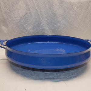 May include: A bright blue enamel casserole dish with two curved handles. The dish has a rounded shape and a glossy finish. This cookware is suitable for baking and serving.