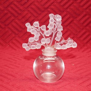 May include: A clear glass perfume bottle featuring a spherical base and a decorative stopper shaped like a cluster of frosted lily of the valley flowers. The stopper is intricately detailed with multiple small, rounded, opaque floral buds.