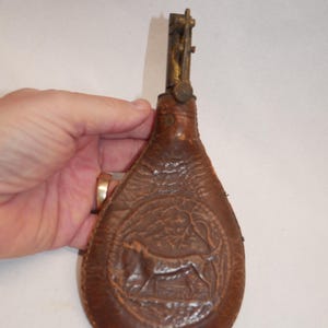Könnte beinhalten: Eine braune Ledertasche mit Messingverschluss. Die Tasche hat ein geprägtes Design eines Hundes in einer Landschaft. Die Tasche wird von einer Hand gehalten.