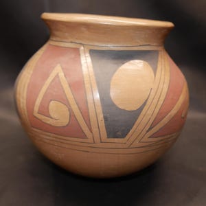May include: A rounded ceramic pot with a wide mouth and a smooth tan base. The exterior features geometric patterns in muted red and dark gray, including triangles, circles, and spiral shapes outlined with thin black lines across the surface.