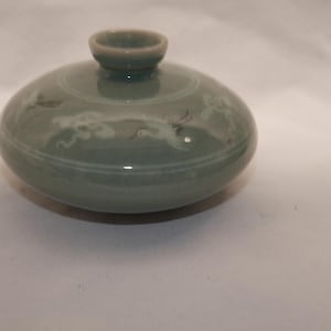 May include: A small, round, celadon-colored ceramic vessel with a short, narrow neck and a wide, flattened body. The surface features delicate, inlaid white designs of flying cranes and stylized clouds encircling the upper portion.