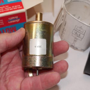 Może przedstawiać: Cylindryczny metalowy silnik elektryczny z wykończeniem w kolorze mosiądzu oraz małym srebrnym wałem wystającym z góry i dołu. Biała prostokątna etykieta na boku obudowy silnika zawiera czarny nadruk z tekstem 6 VDC.