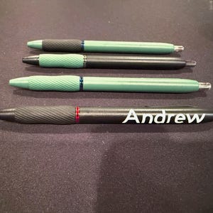 May include: Four pens are displayed on a dark surface. Three pens have green barrels, while one is black. The black pen has the name "Andrew" printed in white. All pens have a textured grip and silver accents.