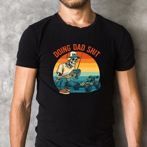 Könnte beinhalten: Schwarzes T-Shirt mit einer Grafik eines Skeletts, das einen "DAD"-Hut trägt und ein ferngesteuertes Auto bedient. Das Design enthält den Text "DOING DAD SHIT" in einem kreisförmigen Design mit einem Sonnenuntergangshintergrund. Das Skelett trägt ein weißes Hemd und blaue Jeans.