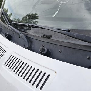 May include: A white vehicle hood featuring two black plastic air intake vents, a black cowl panel, and two black windshield wiper blades positioned against the lower section of the front windshield glass.