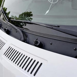 以下が含まれることがあります： 白い車両のボンネットには、通気グリルが組み込まれた黒いプラスチック製のカウルパネル、2本の黒いワイパーアームがあり、フロントガラスには白い充電ケーブルがガラス面に沿って置かれているのが見えます。