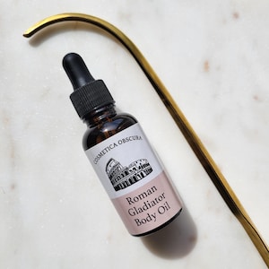 May include: A brown glass bottle with a black dropper, labeled "Cosmetica Obscura Roman Gladiator Body Oil." The label features a drawing of the Colosseum. A gold-colored, curved metal object is in the background. The bottle is on a white marble surface.