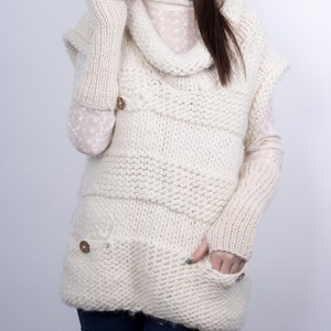 May include: A white knitted sweater with a cowl neck and a large pocket on the front. The sweater has a cable knit pattern and is worn with a white knitted hat.