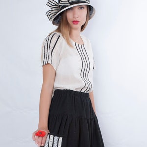 Op de afbeelding: Een vrouw draagt een zwart-wit gestreept blouse, een zwarte rok met witte bies en een zwart-witte hoed. Ze houdt een rode bloem in haar hand.