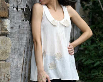 May include: A woman wearing a white sleeveless top with a lace trim and black pants stands next to a weathered wooden wall. She is looking down and has her hair pulled back.