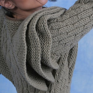 May include: A grey knitted sweater with a unique draped design. The sweater is made from a soft, textured yarn and has a relaxed fit.