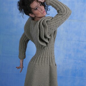 May include: A long-sleeved, knitted sweater dress in a light brown colour. The sweater has a cable knit pattern and a unique design with a large, folded ruffle on the shoulder.