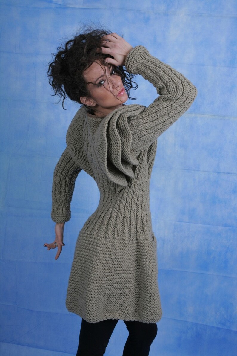 May include: A long-sleeved, knitted sweater dress in a light brown colour. The sweater has a cable knit pattern and a unique design with a large, folded ruffle on the shoulder.