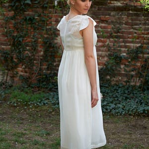 May include: A woman wearing a white, sleeveless dress with a ruffled neckline. She is standing in front of a brick wall with green vines growing on it.