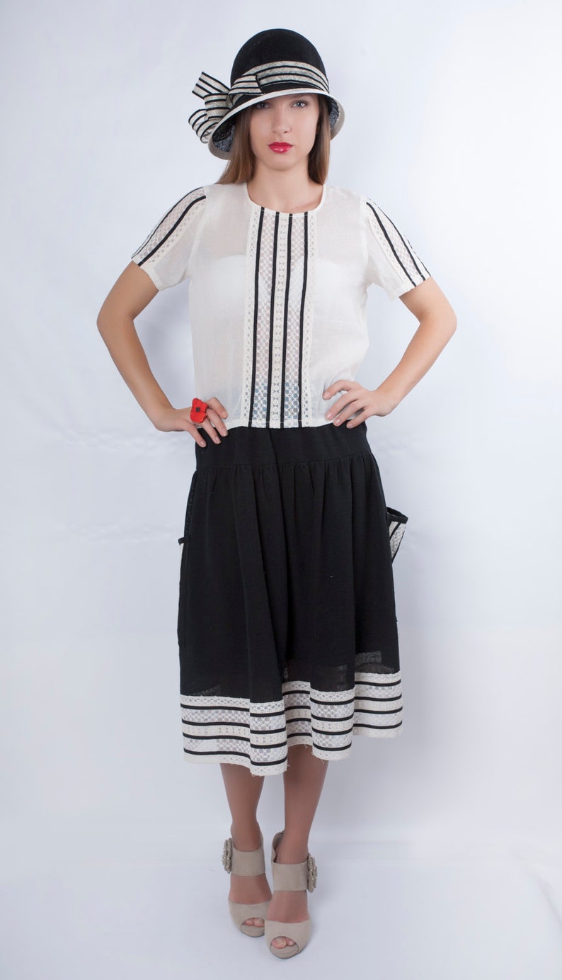 Op de afbeelding: Een vrouw draagt een wit blouse met zwarte strepen en een zwarte rok met witte strepen. Ze draagt ook een zwart-witte hoed met een strik en beige schoenen.