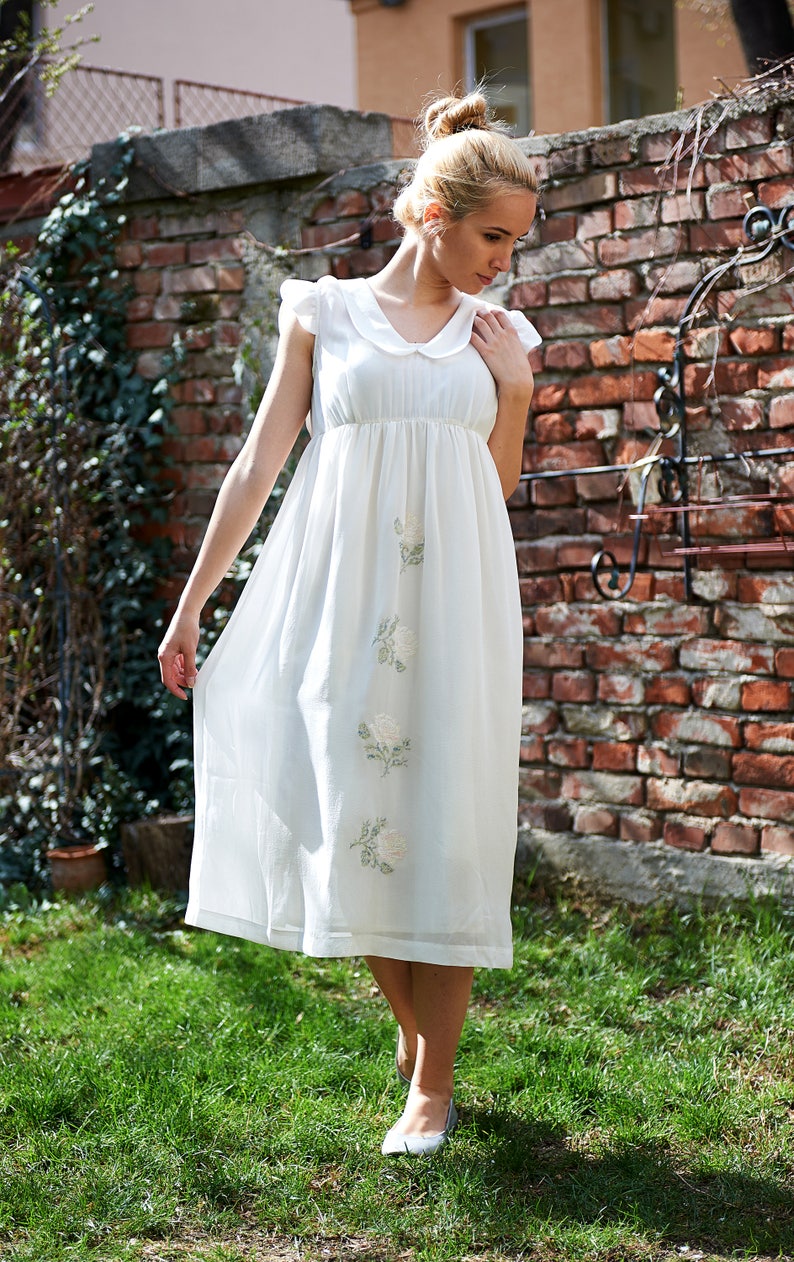 May include: A white cotton dress with a Peter Pan collar and embroidered flowers. The dress has a gathered waist and a flowing skirt.