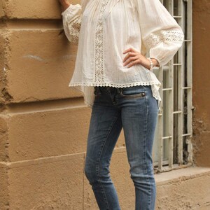 May include: A woman wearing a white crochet top with a lace trim and blue jeans. She is standing next to a wall and has her hand on her hip.