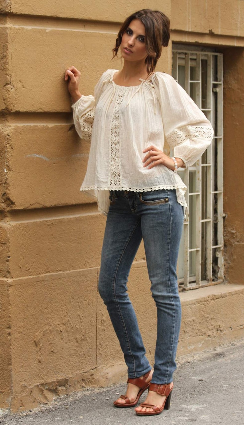 May include: A woman wearing a white crochet top with a lace trim and blue jeans. She is standing next to a wall and has her hand on her hip.