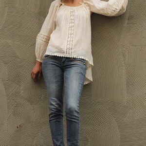 May include: A woman wearing a white crochet top with a lace trim and blue jeans. She is standing in front of a textured wall with a circular pattern.