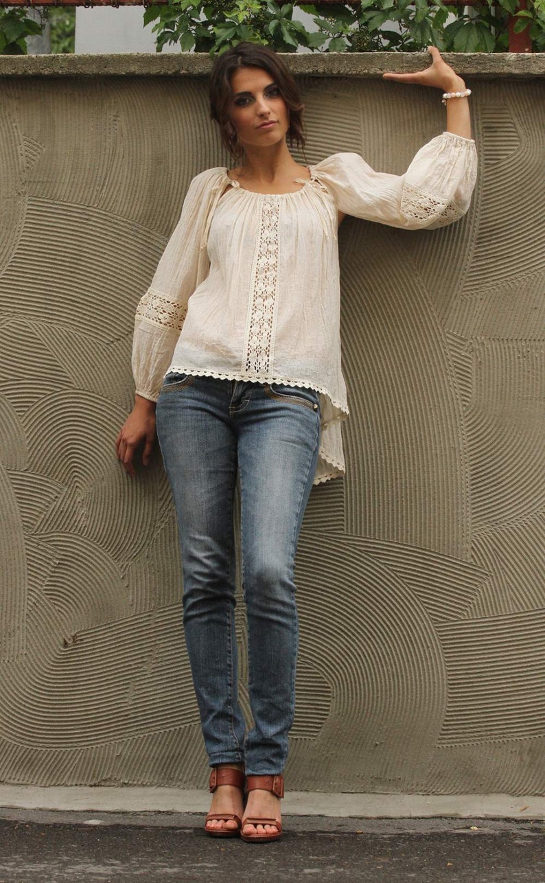 May include: A woman wearing a white crochet top with a lace trim and blue jeans. She is standing in front of a textured wall with a circular pattern.