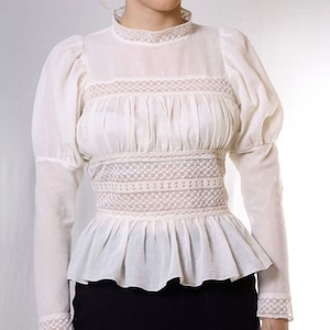 May include: A white cotton blouse with a high neck and long sleeves. The blouse features a lace trim around the neck, waist, and cuffs. The sleeves are puffed at the shoulders and the blouse has a fitted bodice.