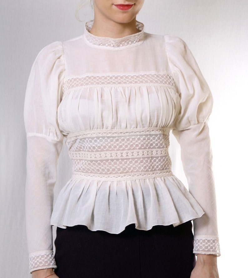 May include: A white cotton blouse with a high neck and long sleeves. The blouse features a lace trim around the neck, waist, and cuffs. The sleeves are puffed at the shoulders and the blouse has a fitted bodice.