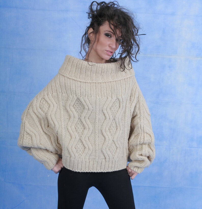 May include: A beige cable knit sweater with a wide, folded turtleneck collar. The sweater has a relaxed fit and long sleeves.