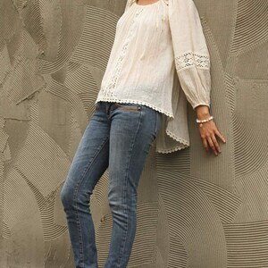 May include: A woman wearing a white crochet top with a lace trim and blue jeans. She is standing in front of a textured wall and wearing brown sandals.