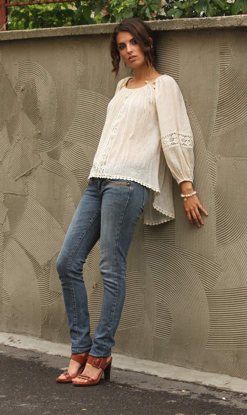 May include: A woman wearing a white crochet top with a lace trim and blue jeans. She is standing in front of a textured wall and wearing brown sandals.