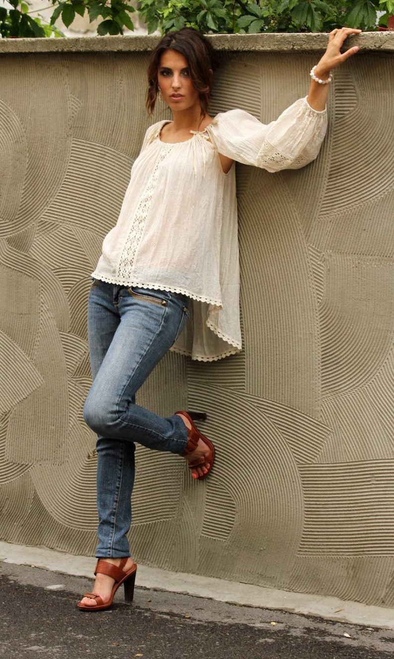 May include: A woman wearing a white crochet top with a lace trim, blue jeans, and brown heeled sandals. She is leaning against a textured wall with a patterned design.