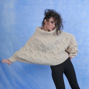May include: A woman wearing a cream-colored, chunky knit sweater with a turtleneck and a cable knit pattern. She is wearing black pants and has her hair pulled back.