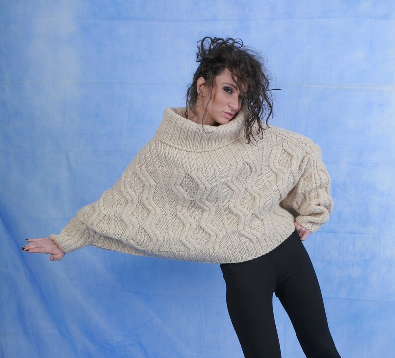 May include: A woman wearing a cream-colored, chunky knit sweater with a turtleneck and a cable knit pattern. She is wearing black pants and has her hair pulled back.