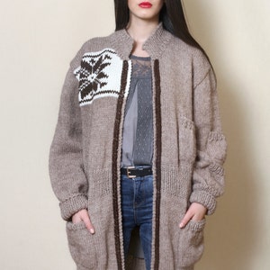 May include: A long, brown knitted cardigan with a white and brown geometric pattern on the front. The cardigan has a zipper closure and two pockets.