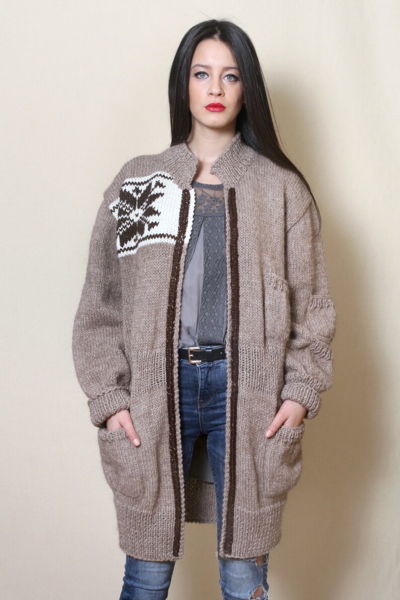 May include: A long, brown knitted cardigan with a white and brown geometric pattern on the front. The cardigan has a zipper closure and two pockets.
