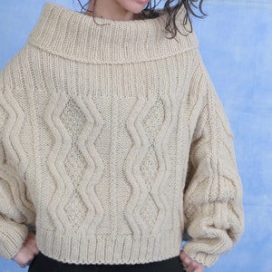 May include: A beige knitted sweater with a cable knit pattern and a wide, folded turtleneck collar.