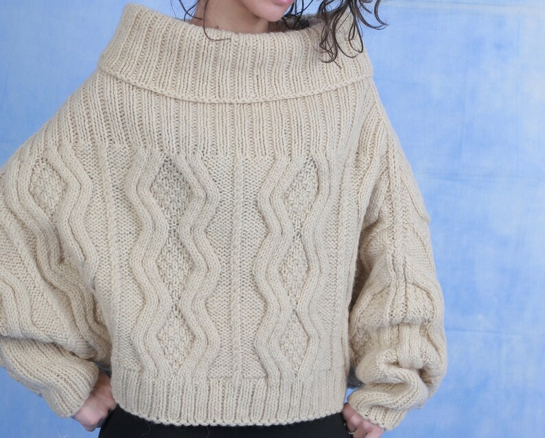 May include: A beige knitted sweater with a cable knit pattern and a wide, folded turtleneck collar.