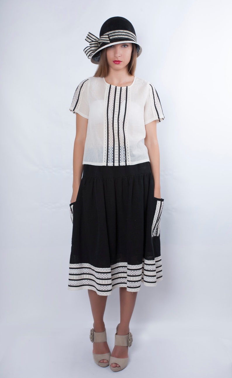Op de afbeelding: Een vrouw draagt een witte blouse met zwarte strepen, een zwarte rok met witte bies en een zwart-witte hoed. Ze draagt beige schoenen met een juweelversiering.