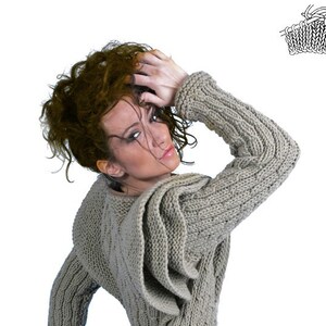 May include: A woman wearing a grey knitted sweater with a large hood. The sweater has a cable knit pattern and a loose fit.