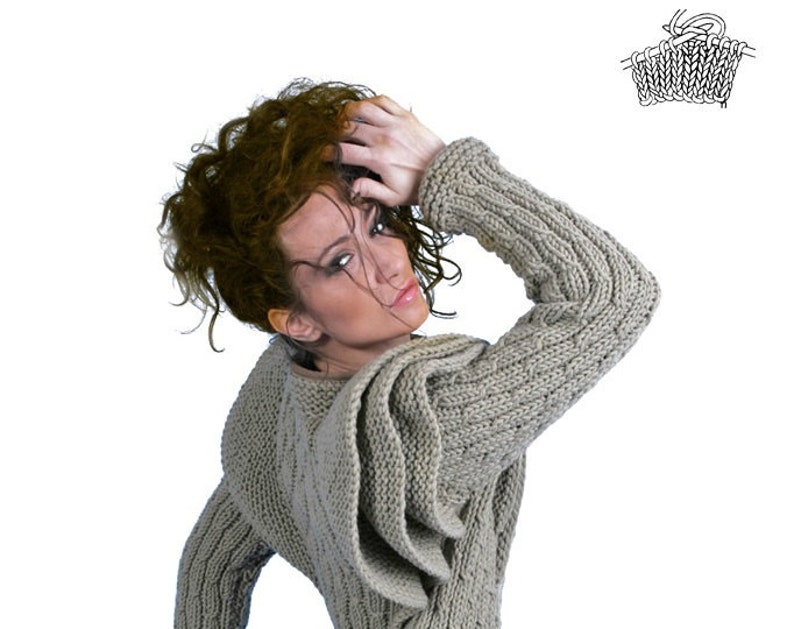 May include: A woman wearing a grey knitted sweater with a large hood. The sweater has a cable knit pattern and a loose fit.