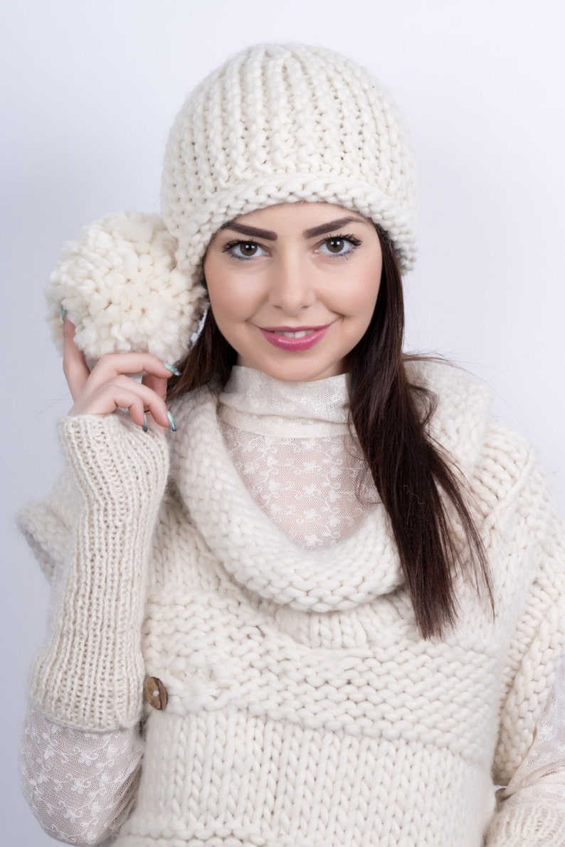 May include: A woman wearing a white knitted hat and a white knitted sweater with a cowl neck. The hat has a pom-pom on top.