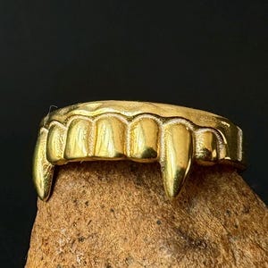 May include: A gold-colored ring shaped like a set of vampire fangs. The ring is displayed on a textured, brown surface against a dark background. The fangs are pointed and detailed.