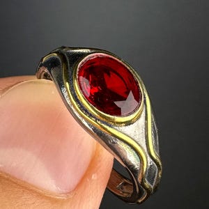May include: A silver ring featuring a large, oval-cut red gemstone. The ring has a gold-coloured band around the gemstone and decorative silver accents. The ring is held between two fingers against a dark background.