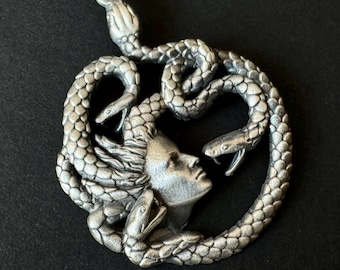 May include: A silver pendant necklace featuring a detailed depiction of Medusa, the Greek mythological figure with snakes for hair. The pendant is circular and shows Medusa's face with snakes intertwined around her head.