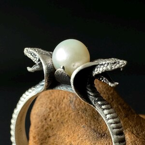 May include: A silver ring featuring two detailed snake heads facing outwards, with a smooth, round white pearl set between them. The band of the ring has a textured, ribbed pattern that mimics the appearance of snake scales, creating a cohesive design.