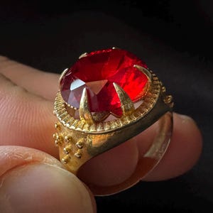 Może przedstawiać: Złoty pierścionek z dużym, czerwonym kamieniem szlachetnym osadzonym w szponach. Pierścionek ma ozdobny wzór wokół kamienia.