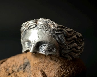 Könnte beinhalten: Ein silberner Ring mit einer detaillierten Skulptur eines Frauenkopfes mit geschlossenen Augen. Der Ring liegt auf einem Stück braunem Stein.