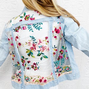 May include: A light blue denim jacket with a sheer floral embroidered panel on the back. The embroidery features pink, red, and green flowers.