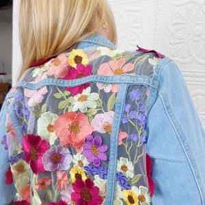 May include: A light blue denim jacket with a sheer floral embroidered overlay. The embroidery features a variety of colorful flowers, including pink, purple, yellow, and green.
