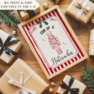May include: A framed print with the text "SON OF A Nutcracker" in a festive design. The print features a red and white striped border and a red illustration of a nutcracker. The image is surrounded by wrapped gifts and holiday decorations.